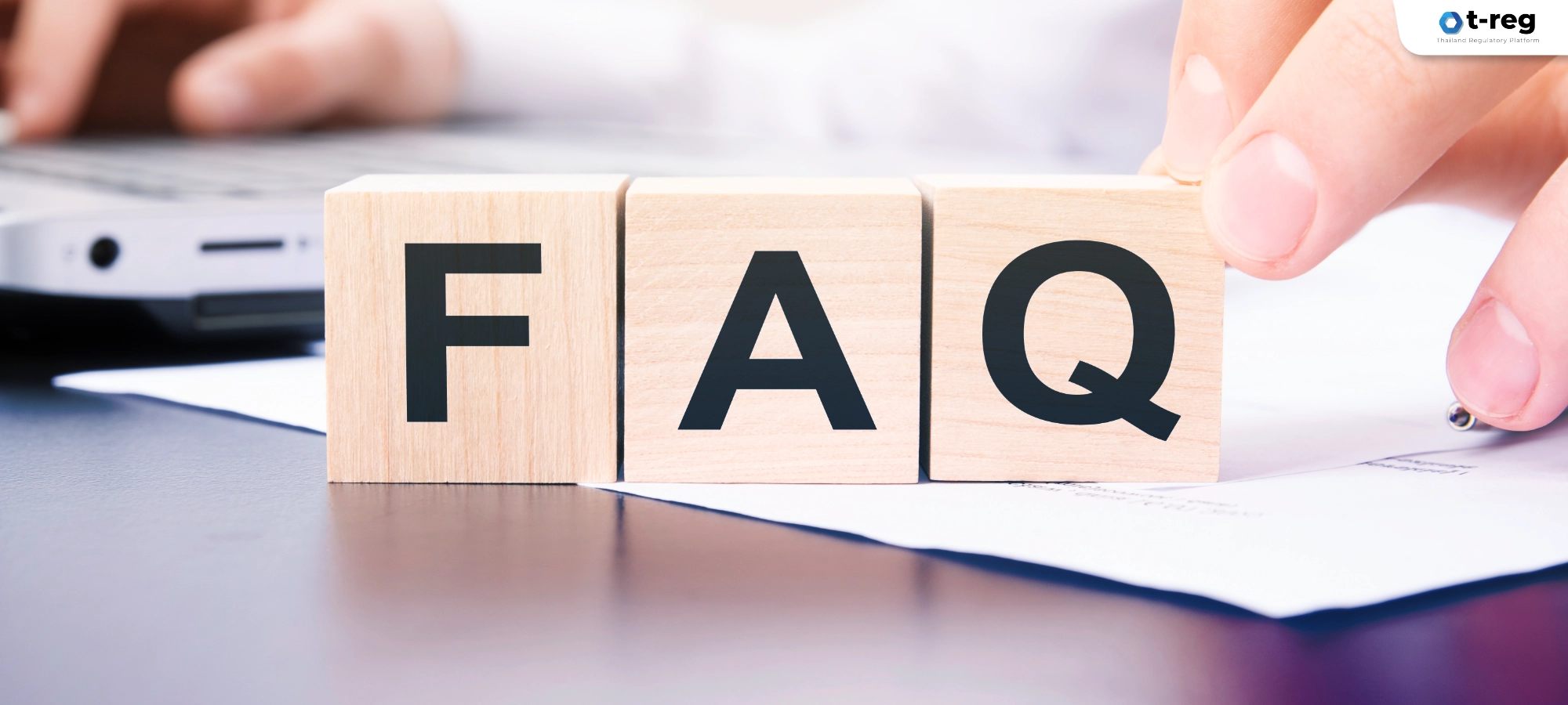 ภาพตัวอักษร FAQ บนบล็อกไม้สื่อถึงคำถามที่พบบ่อย โดยมีมือคนและคอมพิวเตอร์อยู่ด้านหลัง